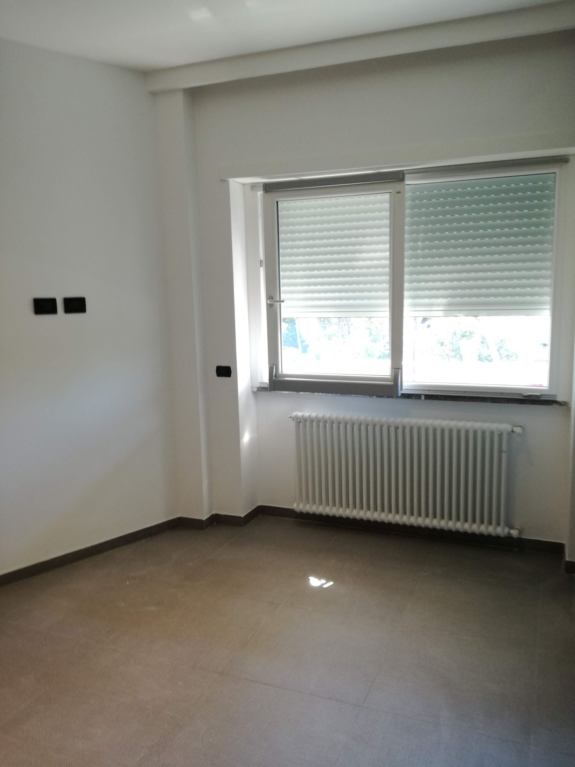 Stanza vuota e non arredata con pareti bianche, pavimento in moquette e una finestra sopra un termosifone.