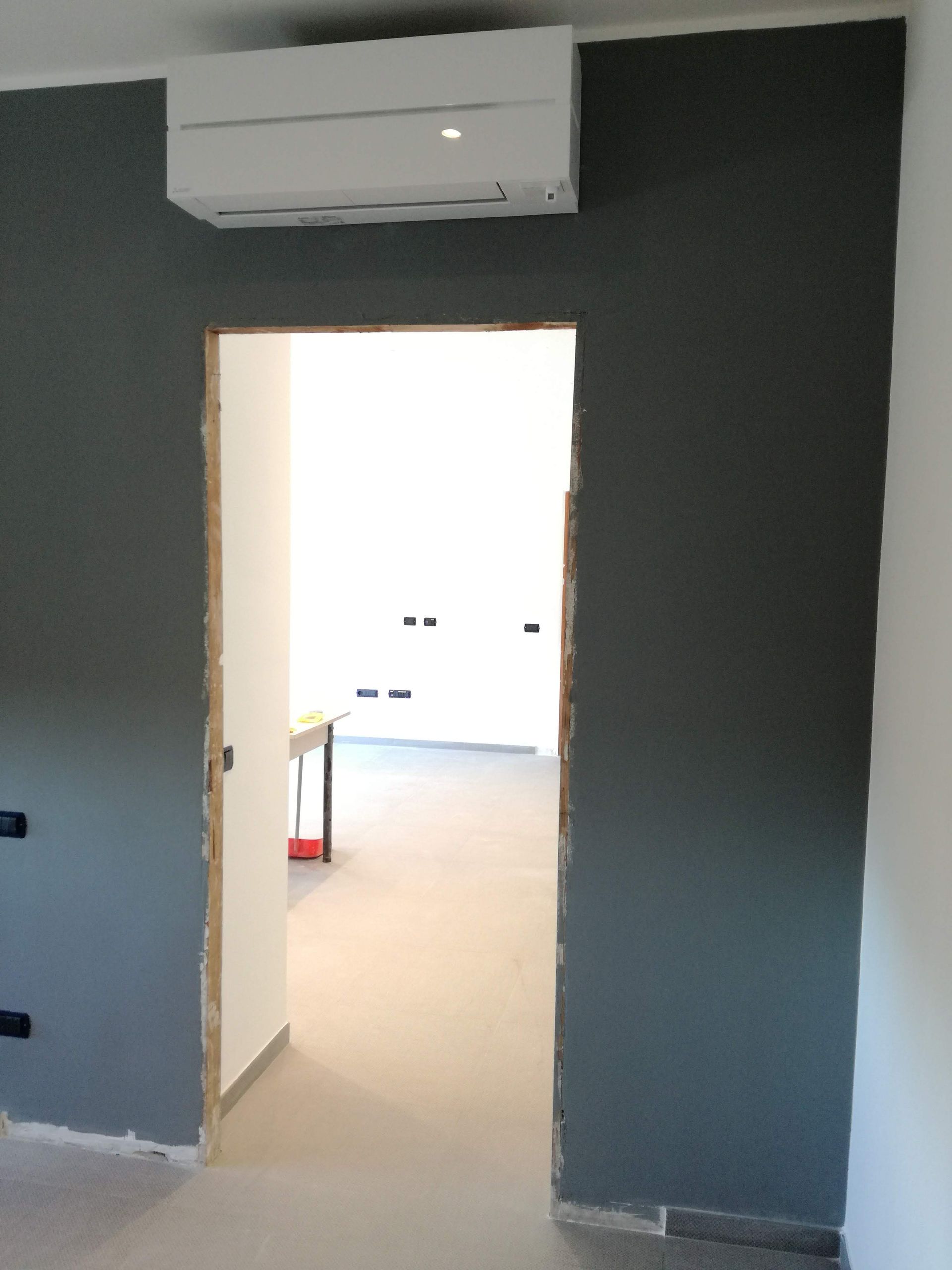 Una porta aperta in una stanza buia, sopra la quale si trova un condizionatore a parete, che conduce a un interno luminoso.