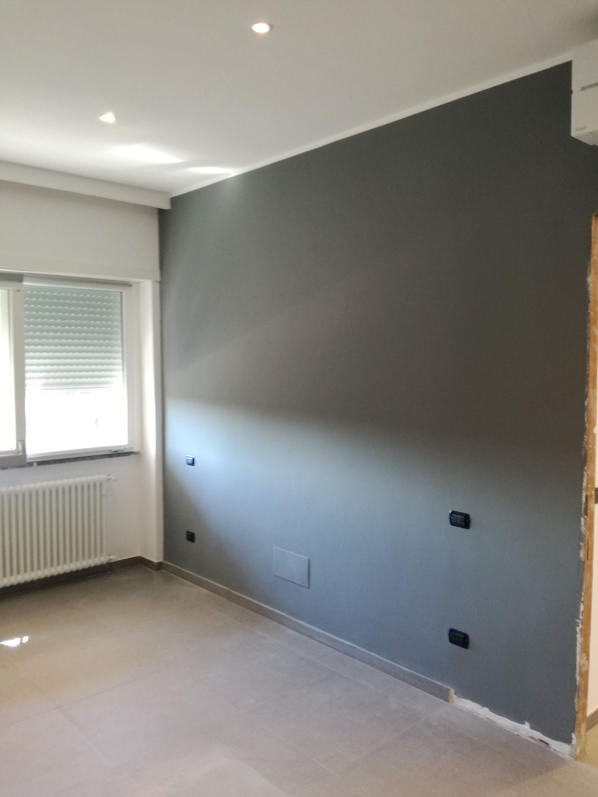 Stanza vuota con parete grigia a contrasto, finestra, termosifone e pavimento rivestito di moquette beige.