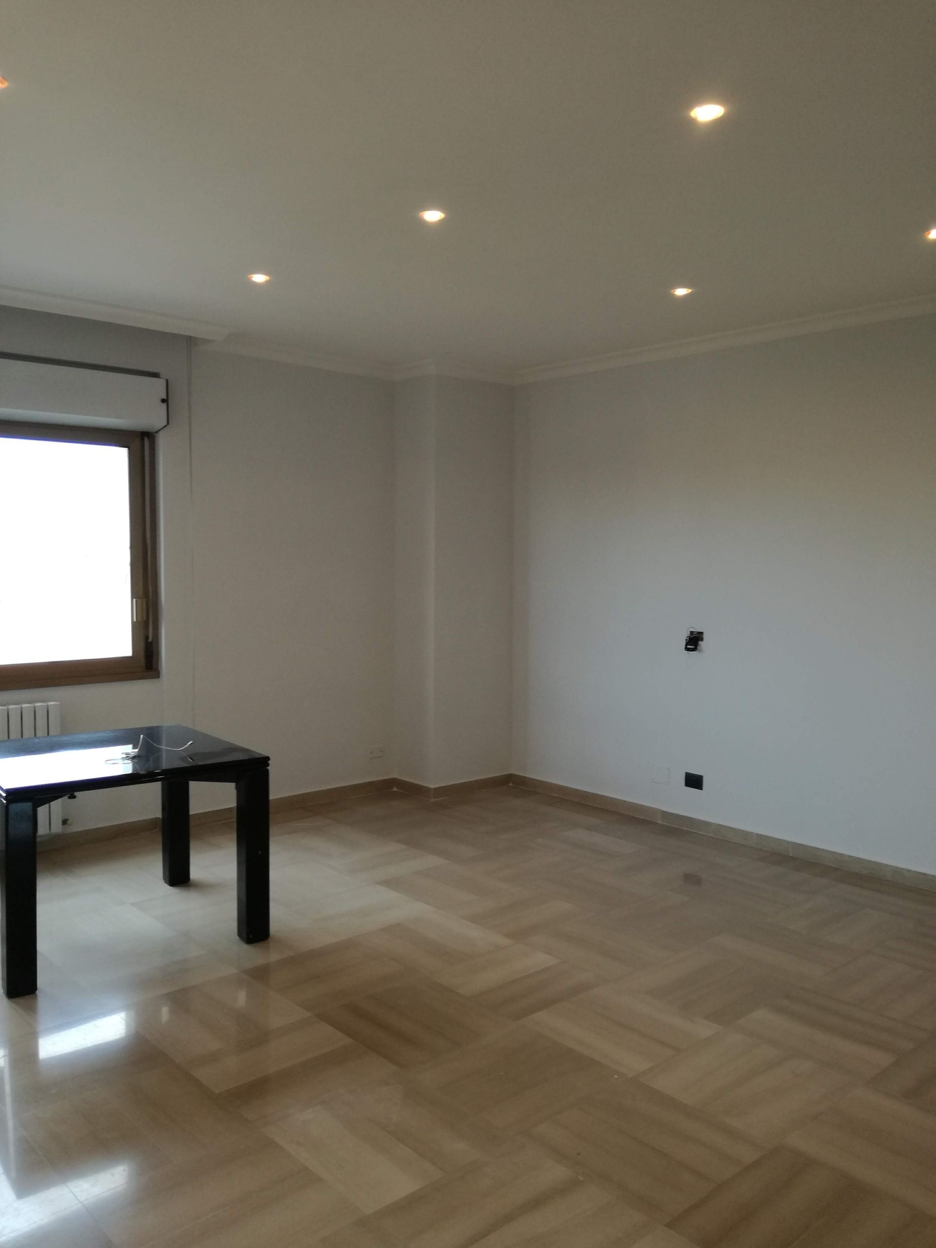 Stanza moderna vuota con pavimento in parquet, pareti bianche, faretti a incasso e una finestra accanto a un tavolo nero.