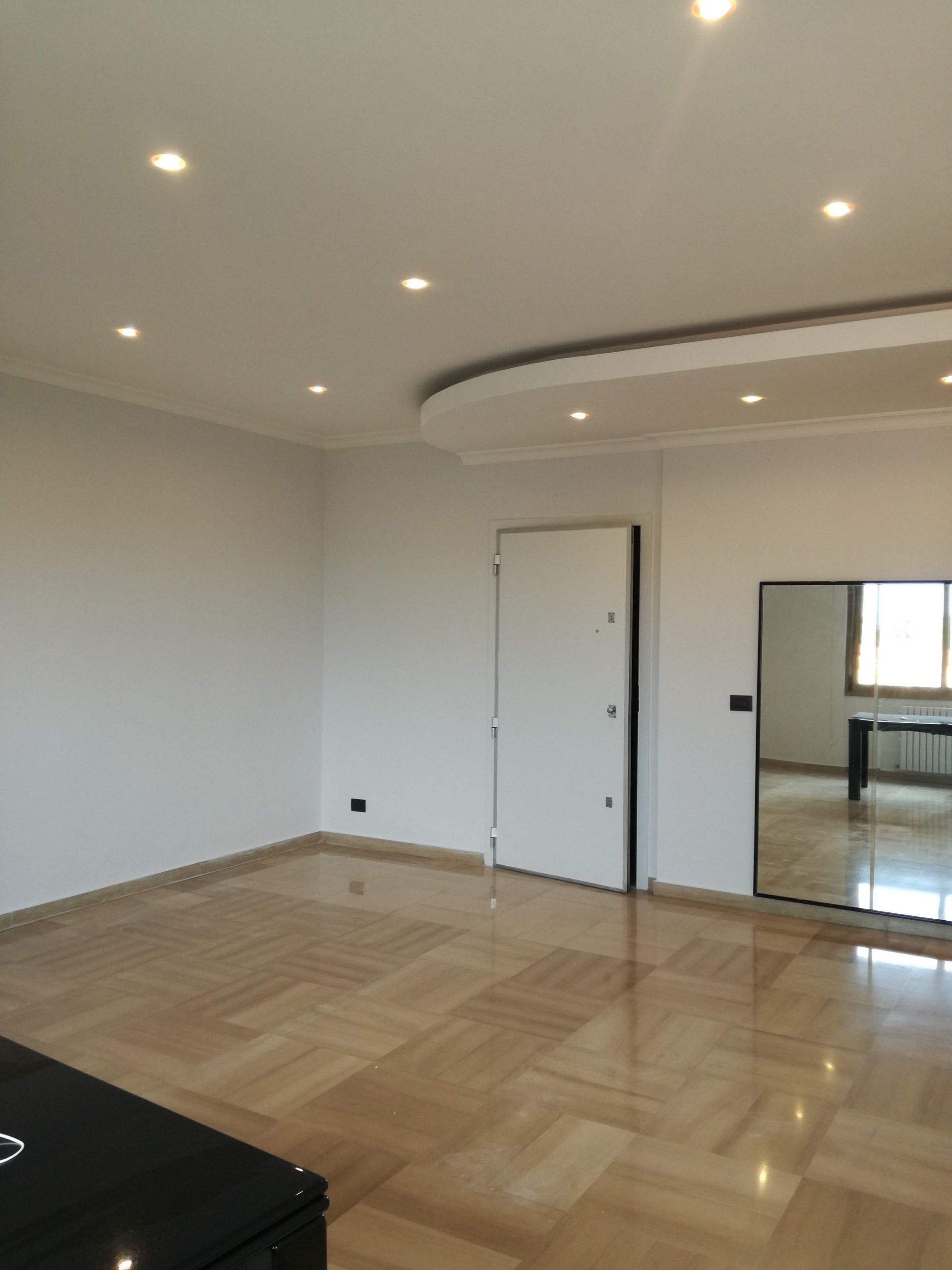 Stanza moderna vuota con pavimento in legno lucido, faretti a incasso sul soffitto, pareti bianche e porta scorrevole a specchio.
