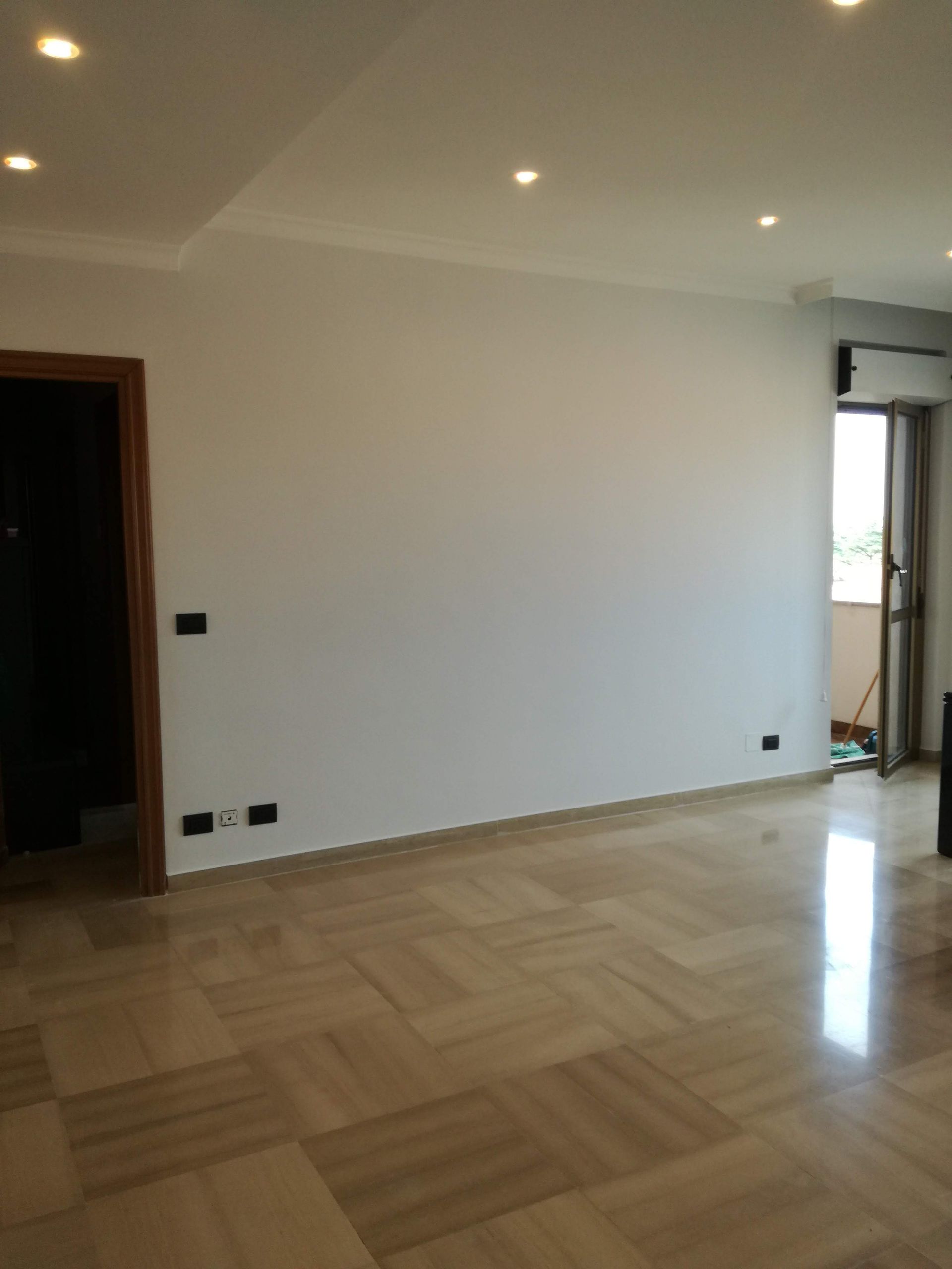 Stanza moderna vuota con pavimento in legno chiaro, pareti bianche, faretti a incasso sul soffitto e porta aperta.
