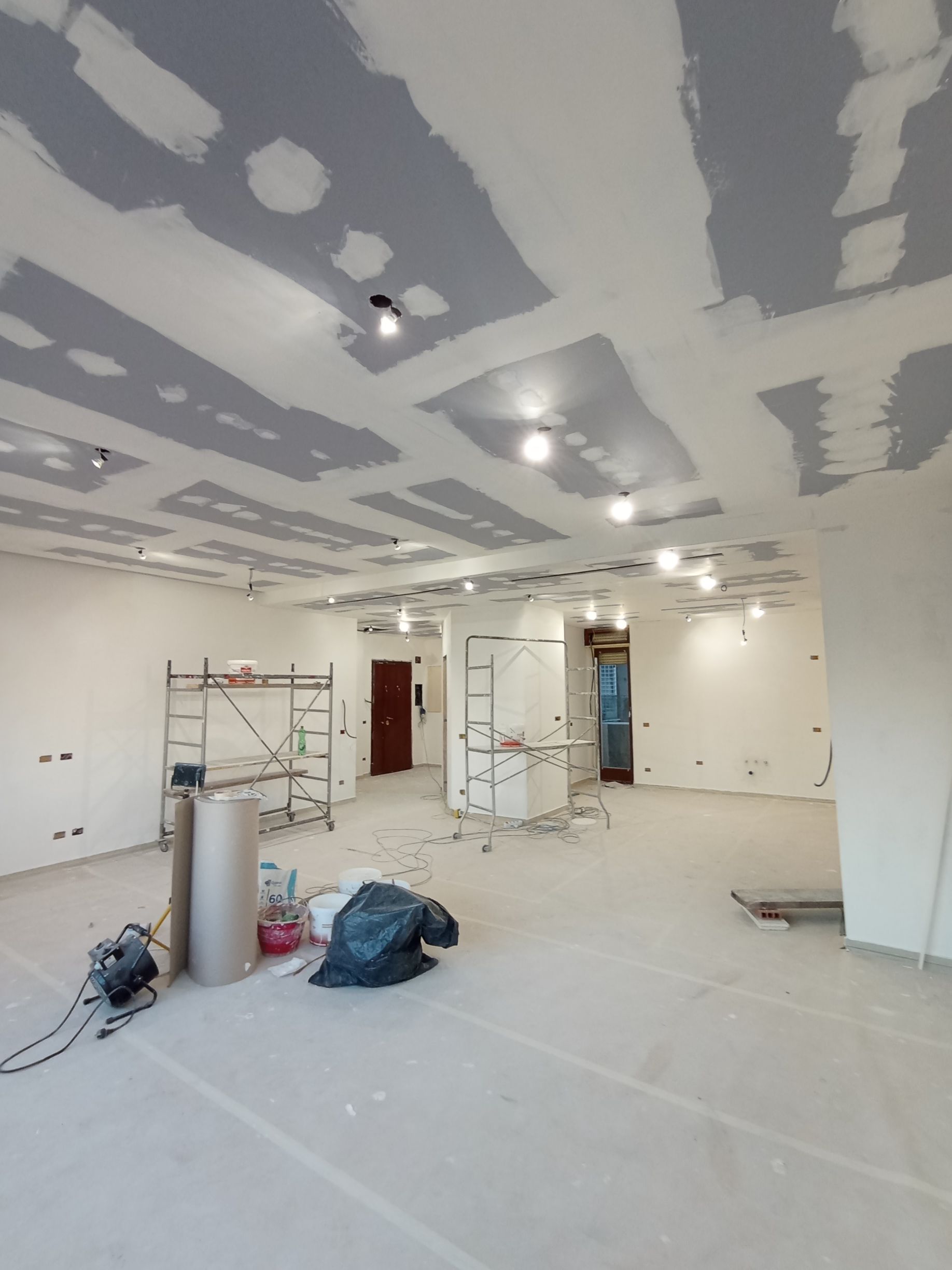 Stanza non finita con rattoppi di cartongesso, impalcature, secchi di vernice e attrezzi da costruzione sul pavimento.
