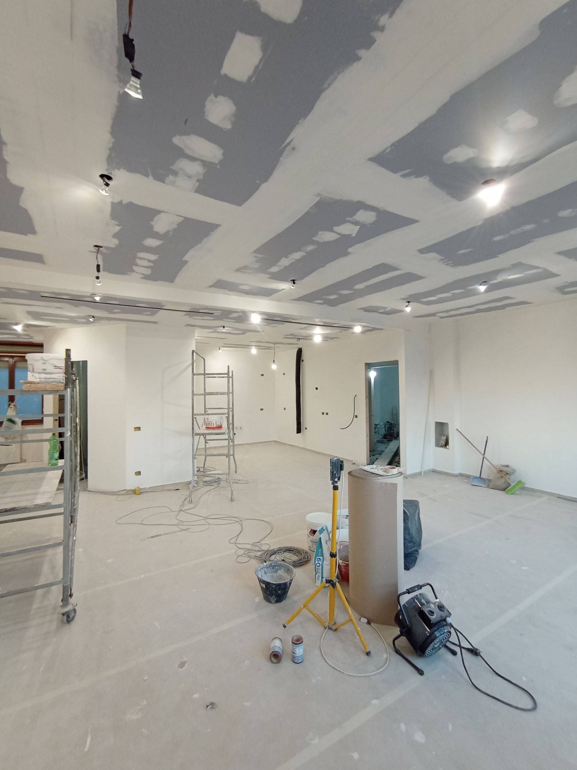 Interno di un ufficio non rifinito, con riparazioni al cartongesso, attrezzi, scale e materiali edili sul pavimento.