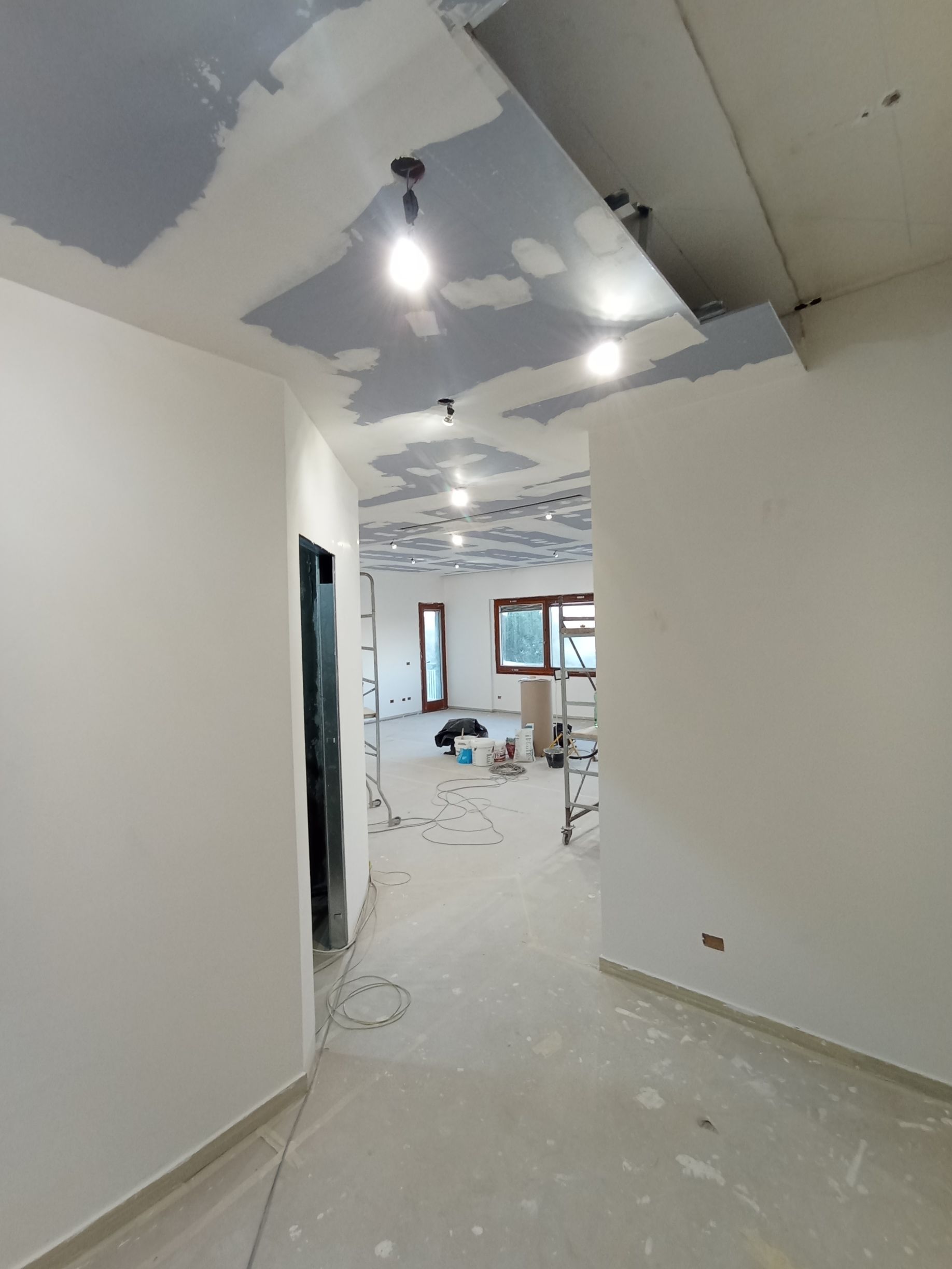 Lavori di ristrutturazione in uno stretto corridoio con soffitto non rifinito, lampade a vista e detriti sul pavimento.