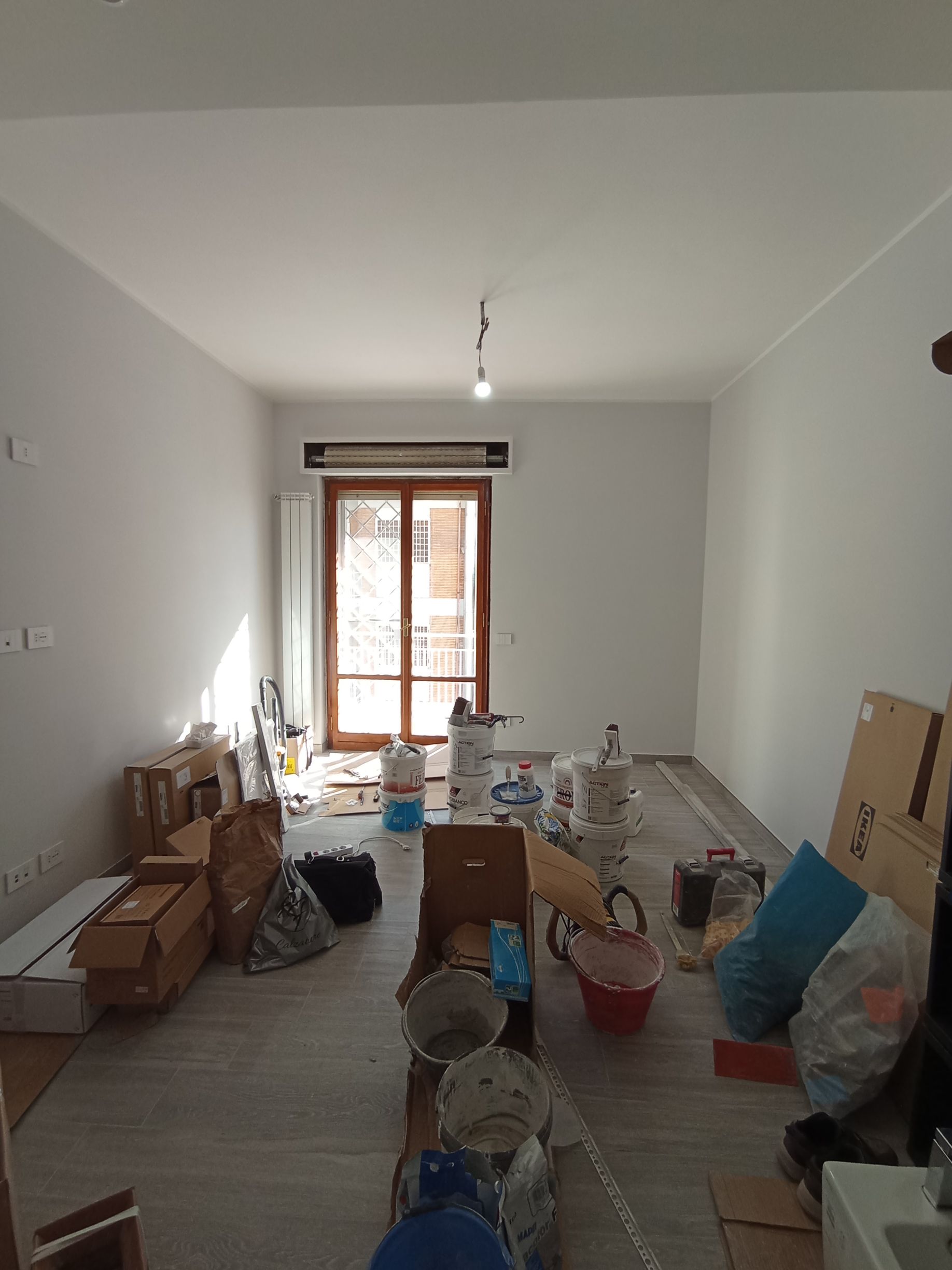 Stanza lunga e stretta in fase di ristrutturazione, con scatole, secchi e attrezzi sparsi sul pavimento.