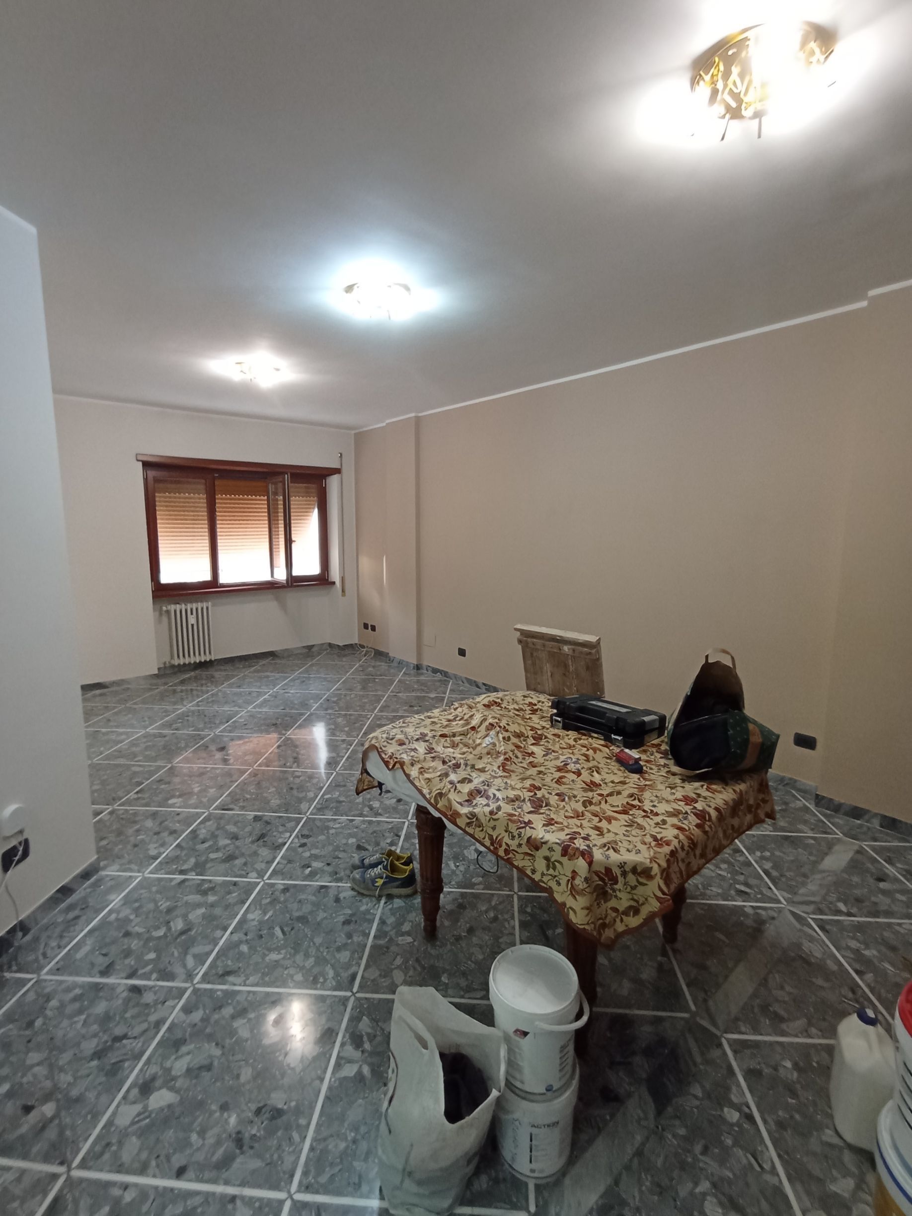 Stanza vuota e non arredata con pavimento piastrellato, luci a soffitto, una finestra e un tavolino con oggetti in primo piano.