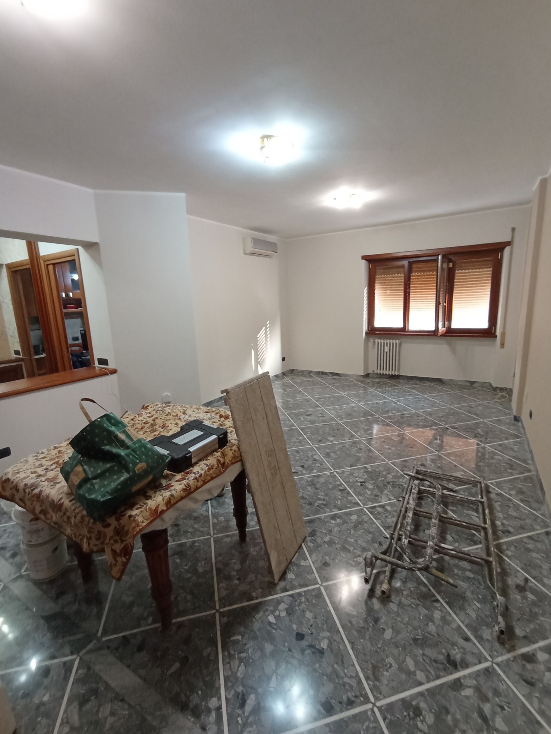 Soggiorno vuoto con pavimento piastrellato, tende alle finestre, tavolo e sgabello vicino al centro.