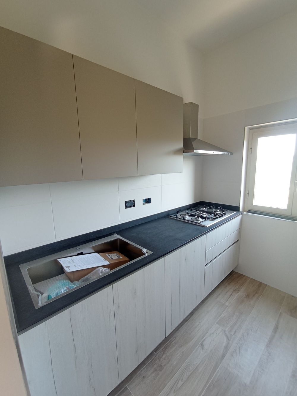 Cucina moderna con mobili in legno chiaro, piano di lavoro nero, piano cottura a gas, lavello e finestra sopra il bancone.