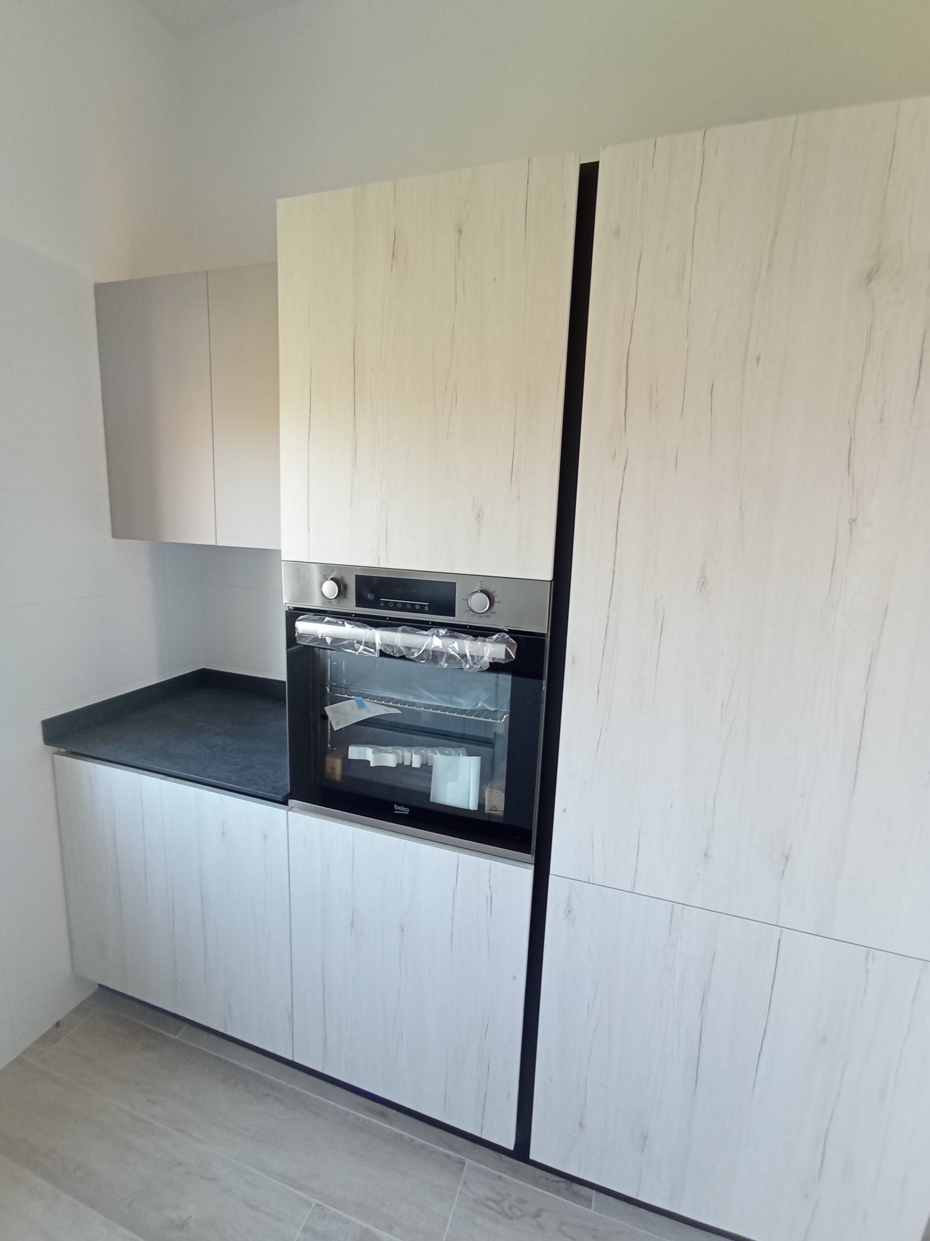 Cucina moderna con mobili in legno chiaro, ripiani neri e forno a incasso.