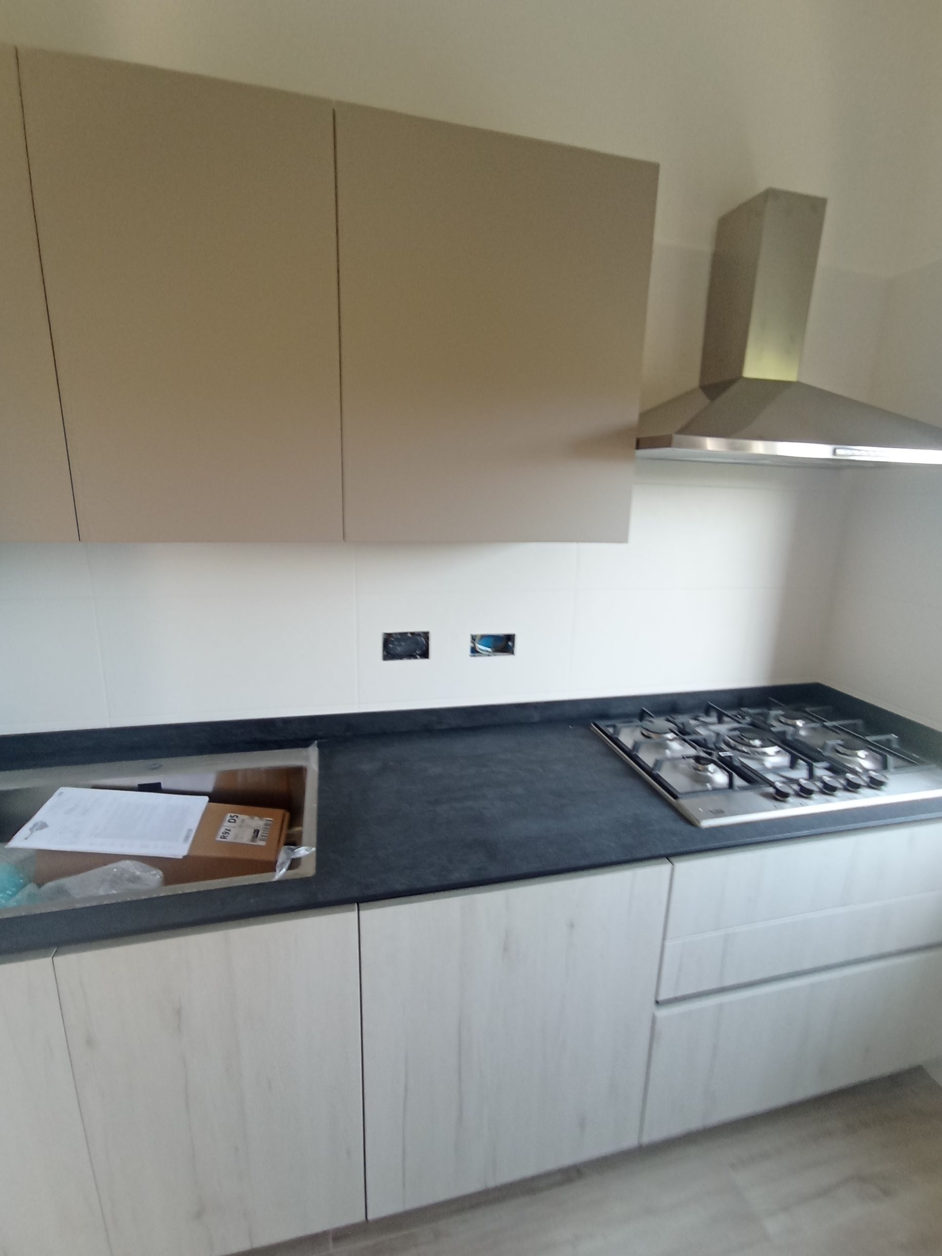 Cucina moderna con mobili beige, piano di lavoro scuro, piano cottura a gas e cappa aspirante in acciaio inox.