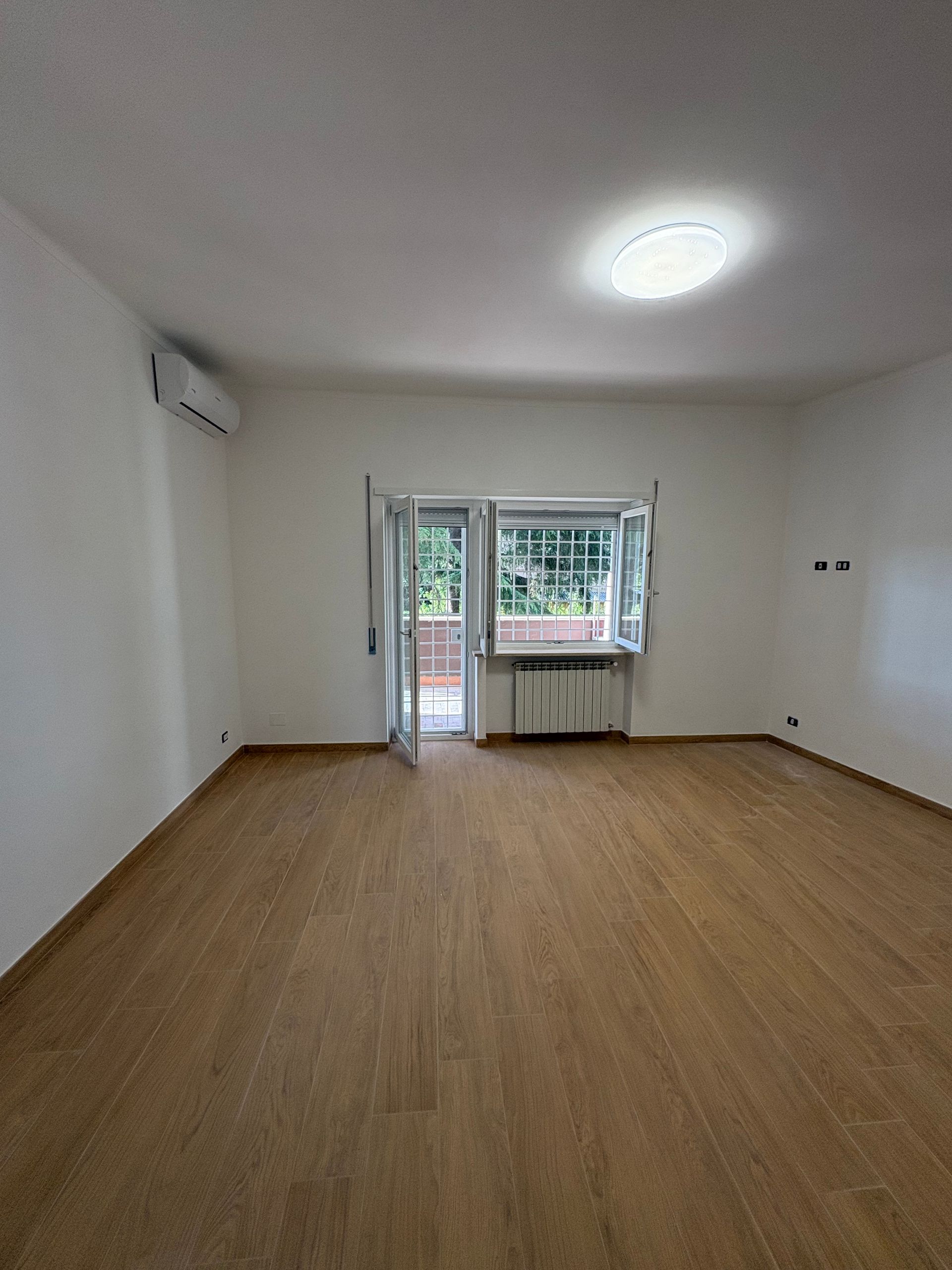 Stanza vuota e non arredata con pavimento in legno, pareti bianche, lampadario a soffitto e porta finestra che dà sul balcone.