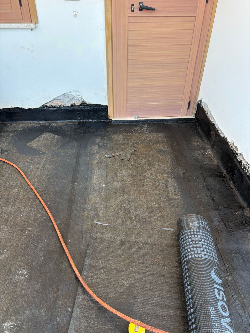 Pavimento appena impermeabilizzato accanto a una porta di legno, con un tubo arancione e una membrana impermeabile stesa a terra.