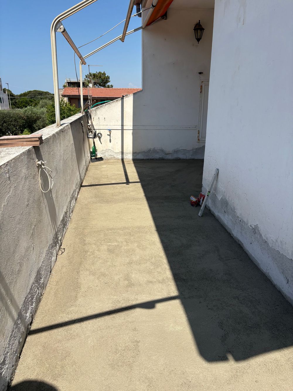 Uno stretto balcone soleggiato accanto a un edificio bianco, con un muretto basso e un'ombra profonda sul pavimento.