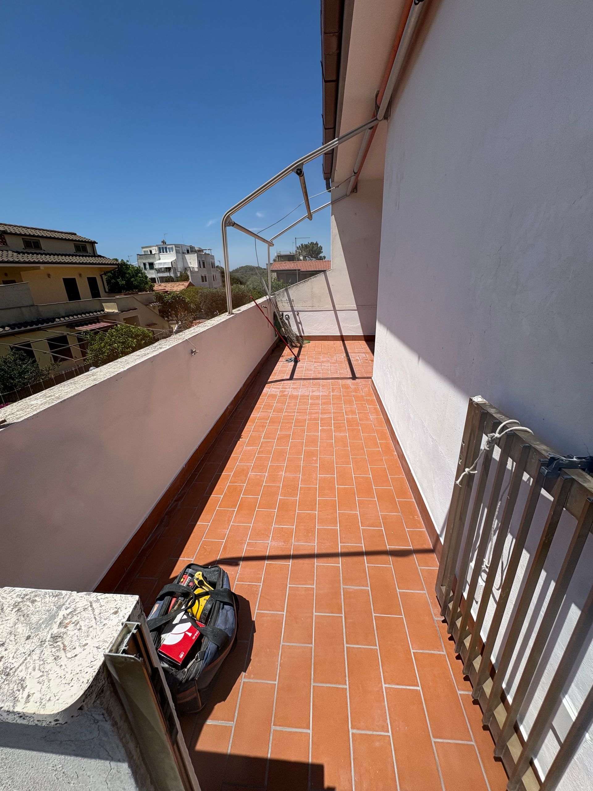 Balcone stretto e soleggiato con piastrelle in terracotta, pareti bianche e una borsa nera sul pavimento.