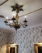 Un lampadario dorato e decorato pende da un soffitto bianco sopra una carta da parati a motivi e opere d'arte incorniciate.