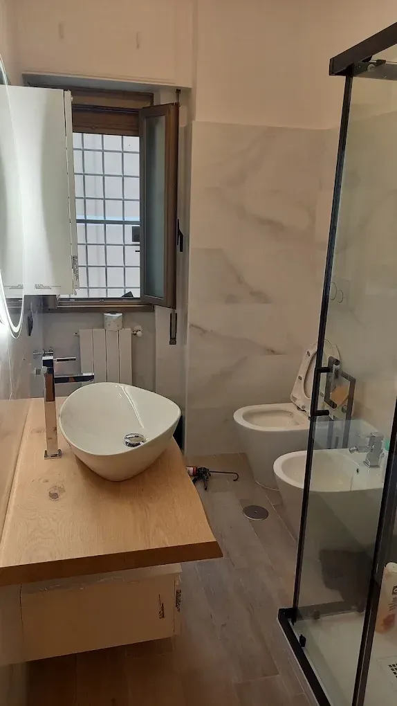 Piccolo bagno con mobiletto in legno, lavabo rotondo, box doccia in vetro e luce a finestra.