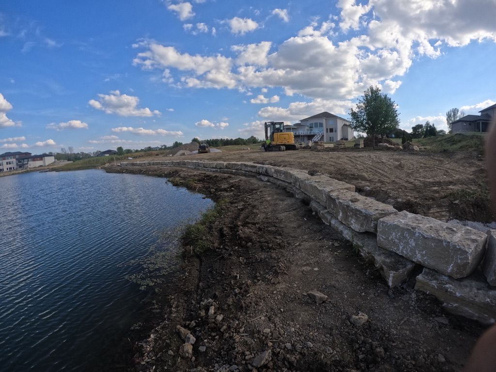 A panoramic view of a construction site next to a body of water, featuring an excavator, a partially built retaining wall
