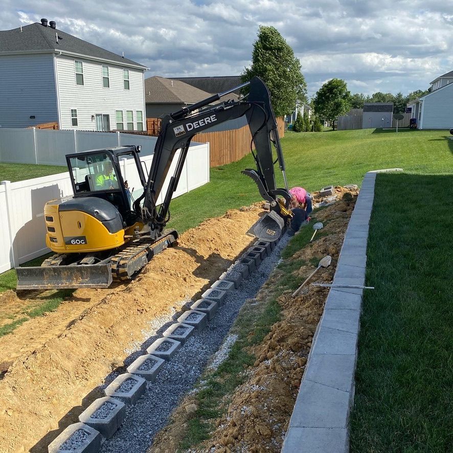 John Deere 60G compact excavator digging a trench in a residential backyard with a worker assisting.