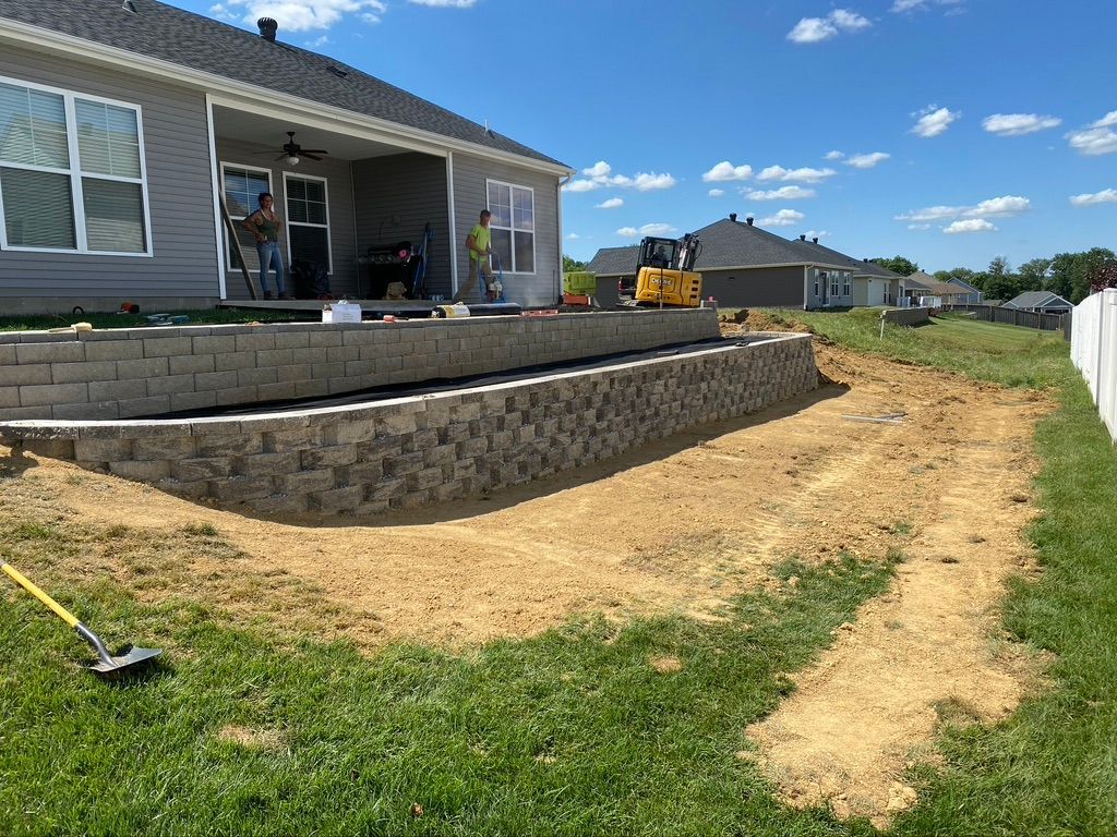 Construction site with a newly built stone retaining wall in front of a house, featuring an excavator and workers