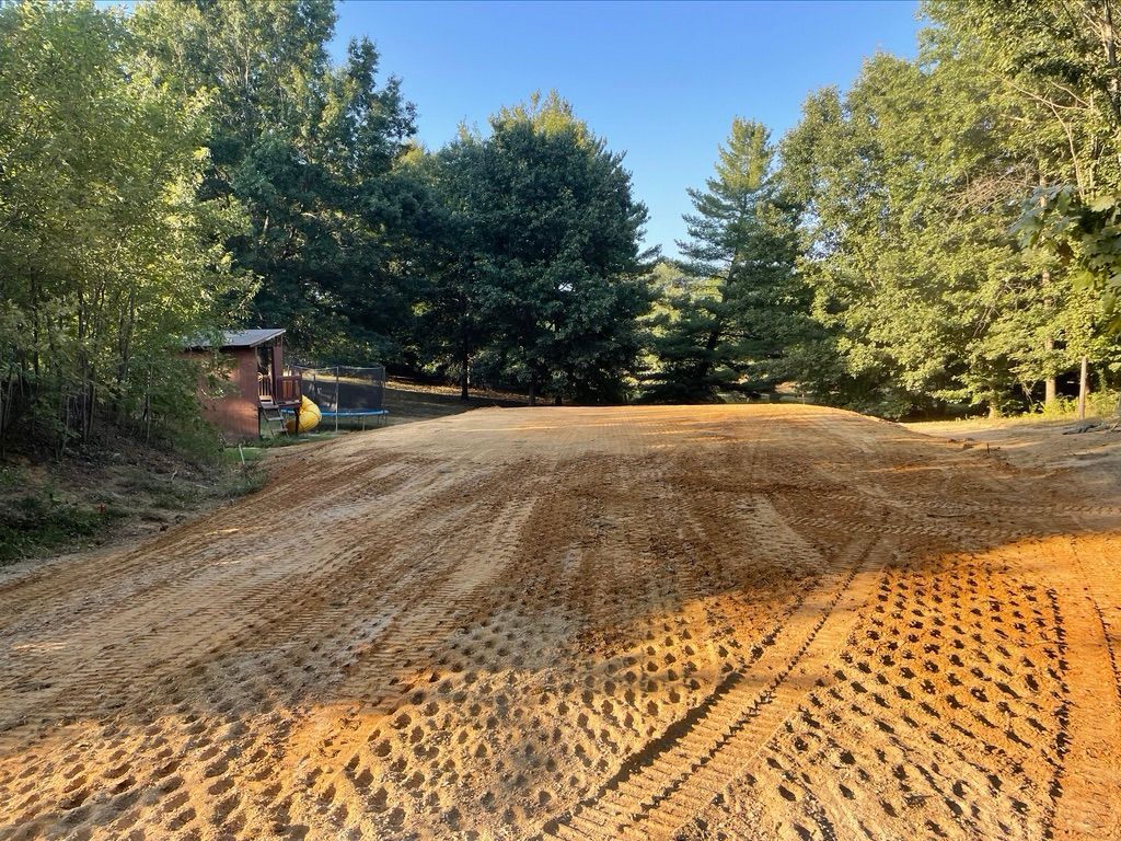 Cleared and leveled building lot in a wooded area with a small shed and trampoline in the background