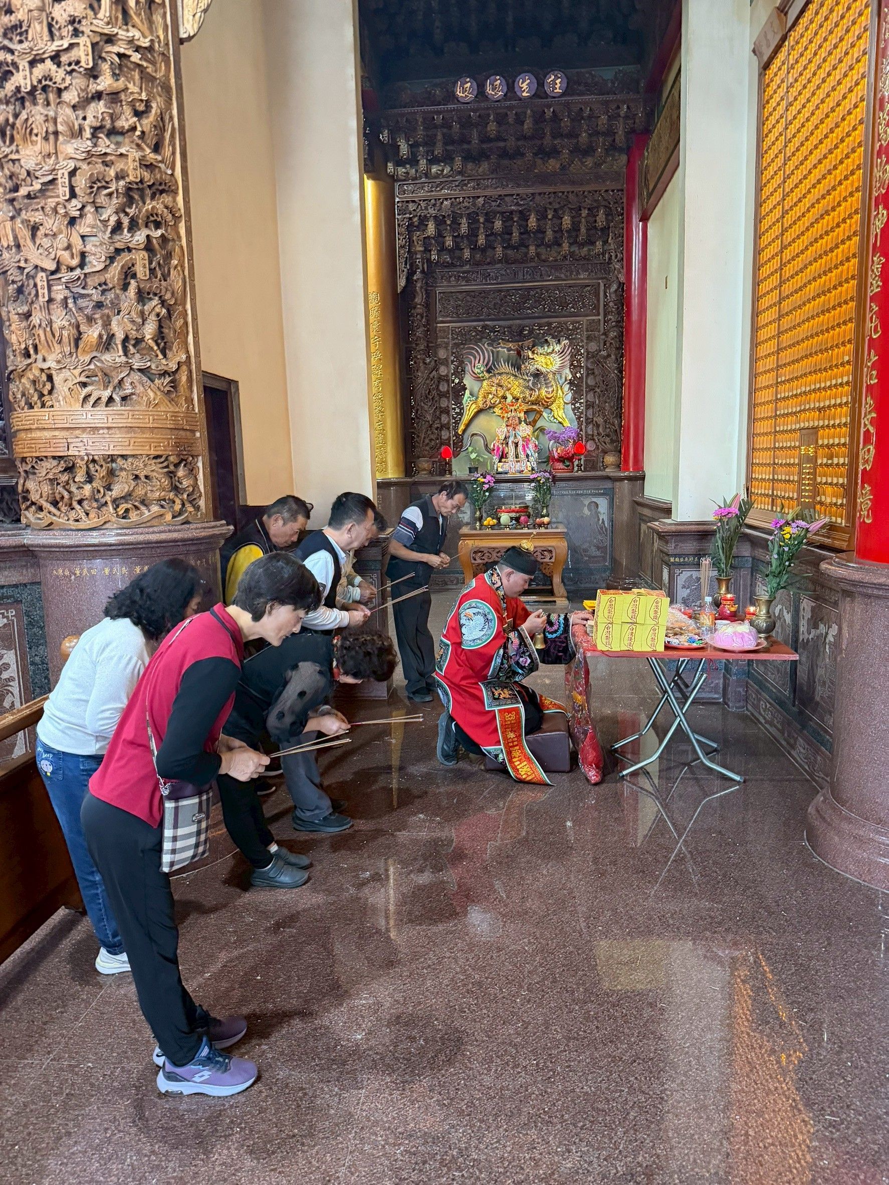 人們在寺廟祭壇前祈禱，背景可見祭品和神像。華麗的柱子和金色裝飾點綴其間。