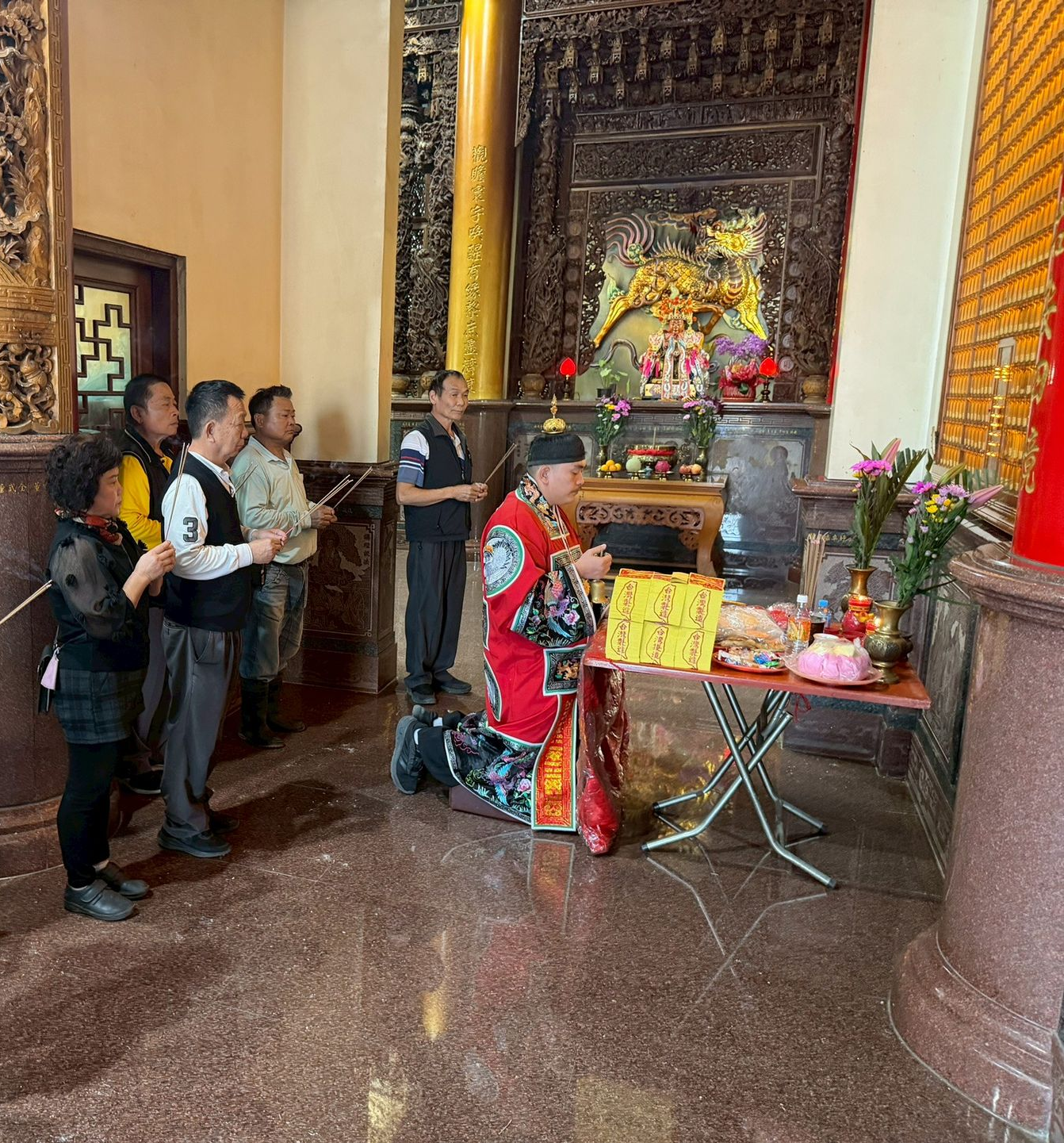 在寺廟場景中，一名身穿紅色長袍的人跪在祭壇前，周圍有其他人。