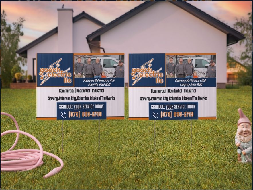 Two yard signs for an electrical company are displayed in a lawn in front of a house, beside a garden hose and a gnome.