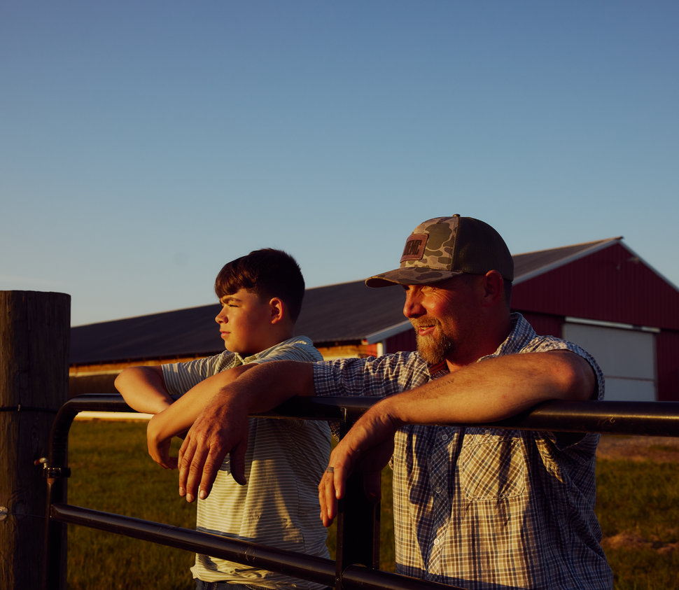 Man with arm around a boy's shoulder walking down a farm path past outbuildings on a sunny day.