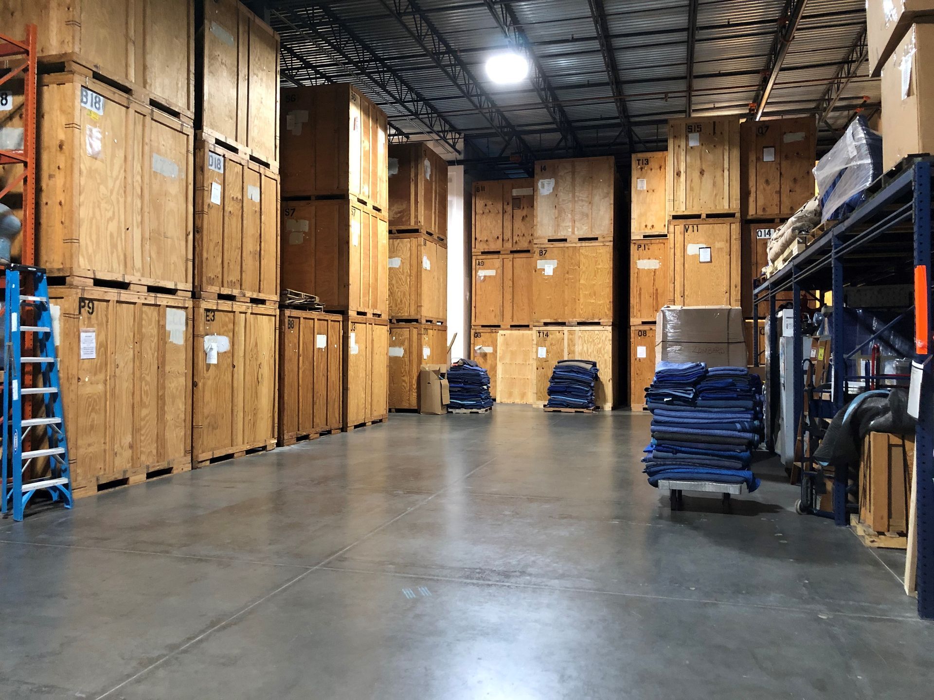 Warehouse interior, rows of large wooden crates stacked high. Gray concrete floor, metal shelving, and blue ladder.