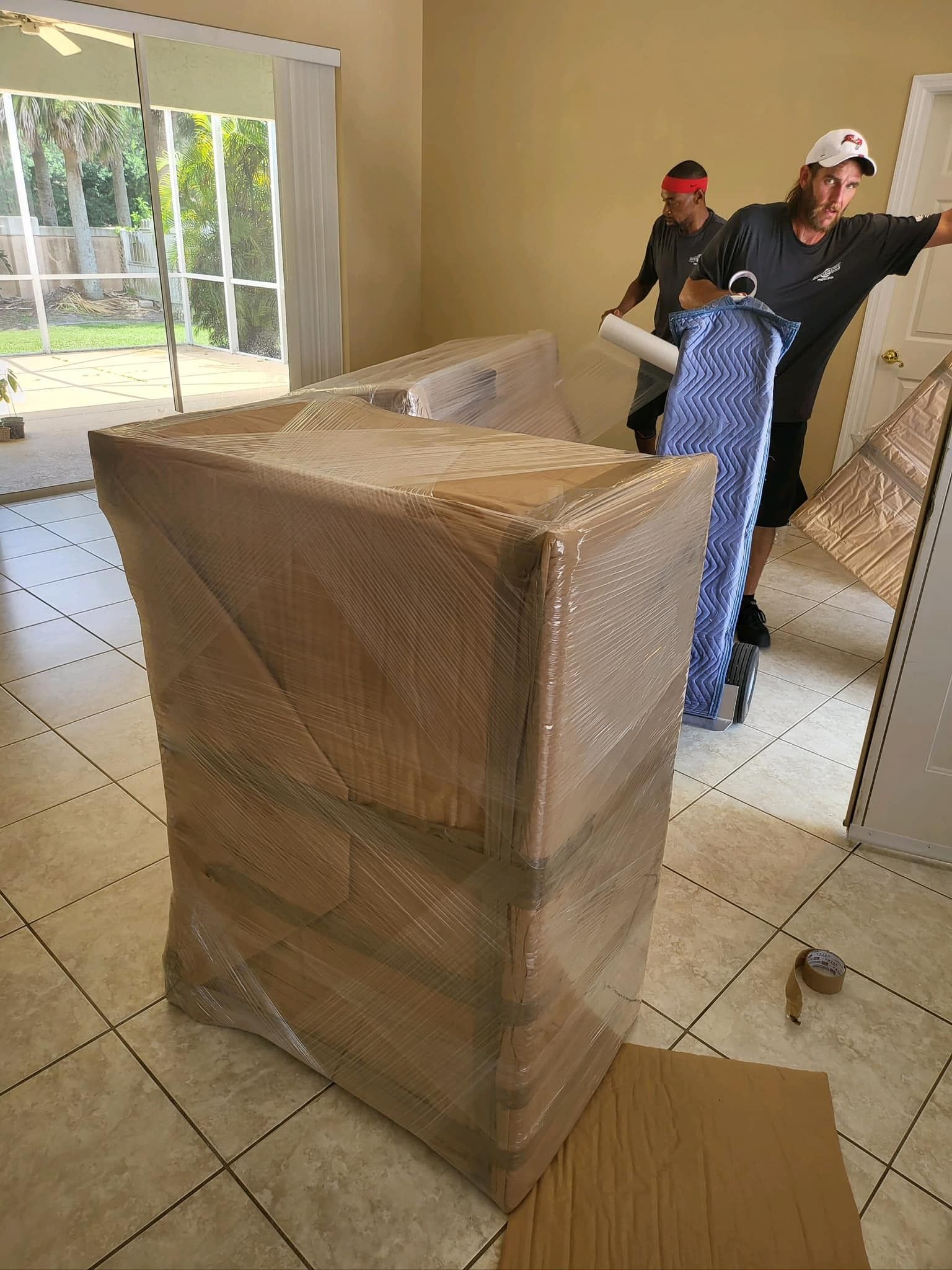 Movers wrapping furniture with plastic in an empty room with large glass doors.