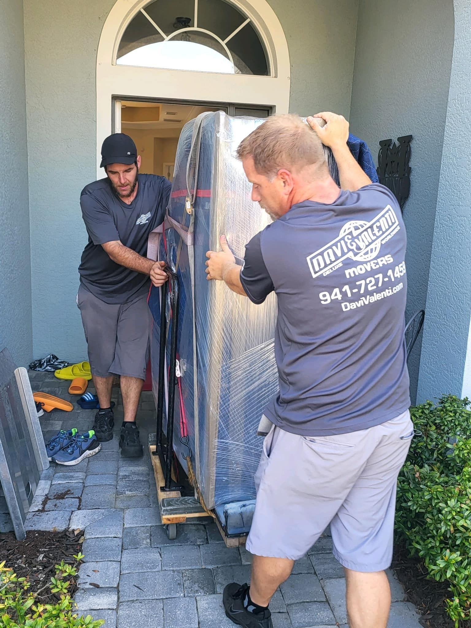 Two movers carrying a large wrapped object out of a house doorway.