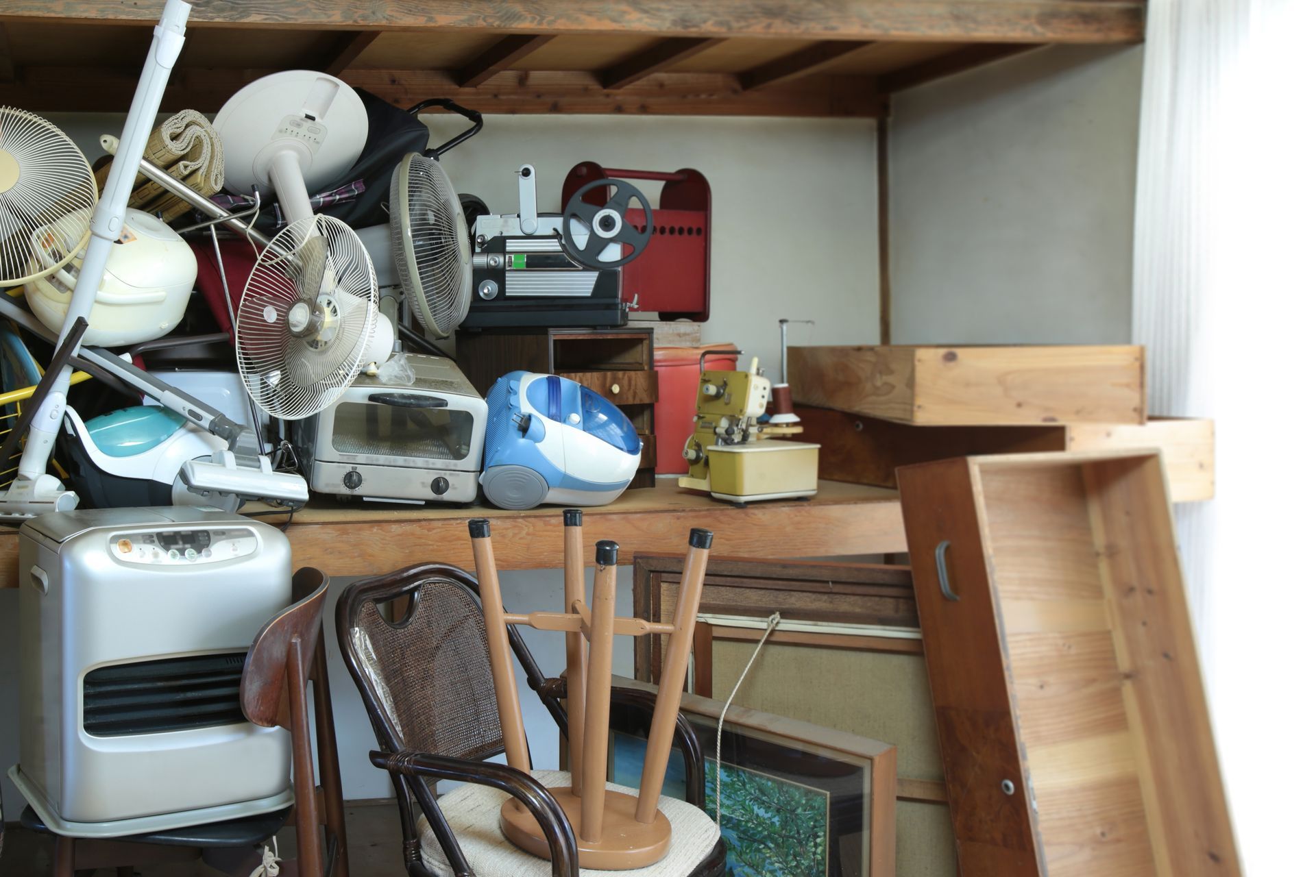 Cluttered storage room with stacked chairs, boxes, lamps, and wooden boards under a low shelf