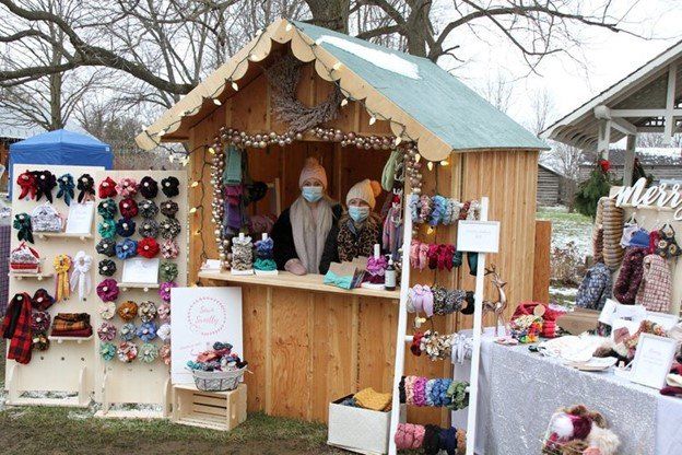 Two people wearing masks and winter clothing stand inside a small wooden chalet, that has Christmas decorations on the outside of it. Various scrunchies and headbands are displayed on tables outside the chalet for people to purchase.