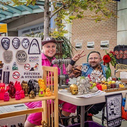 An image of two people making peace signs behind a booth of jacket patches and stickers.