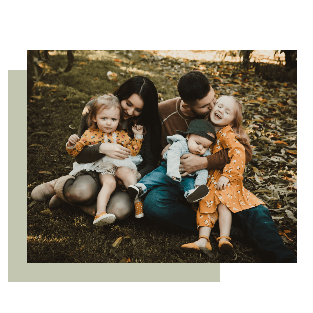 Family of five sits on grass, smiles, and cuddles. Fall colors surround them.