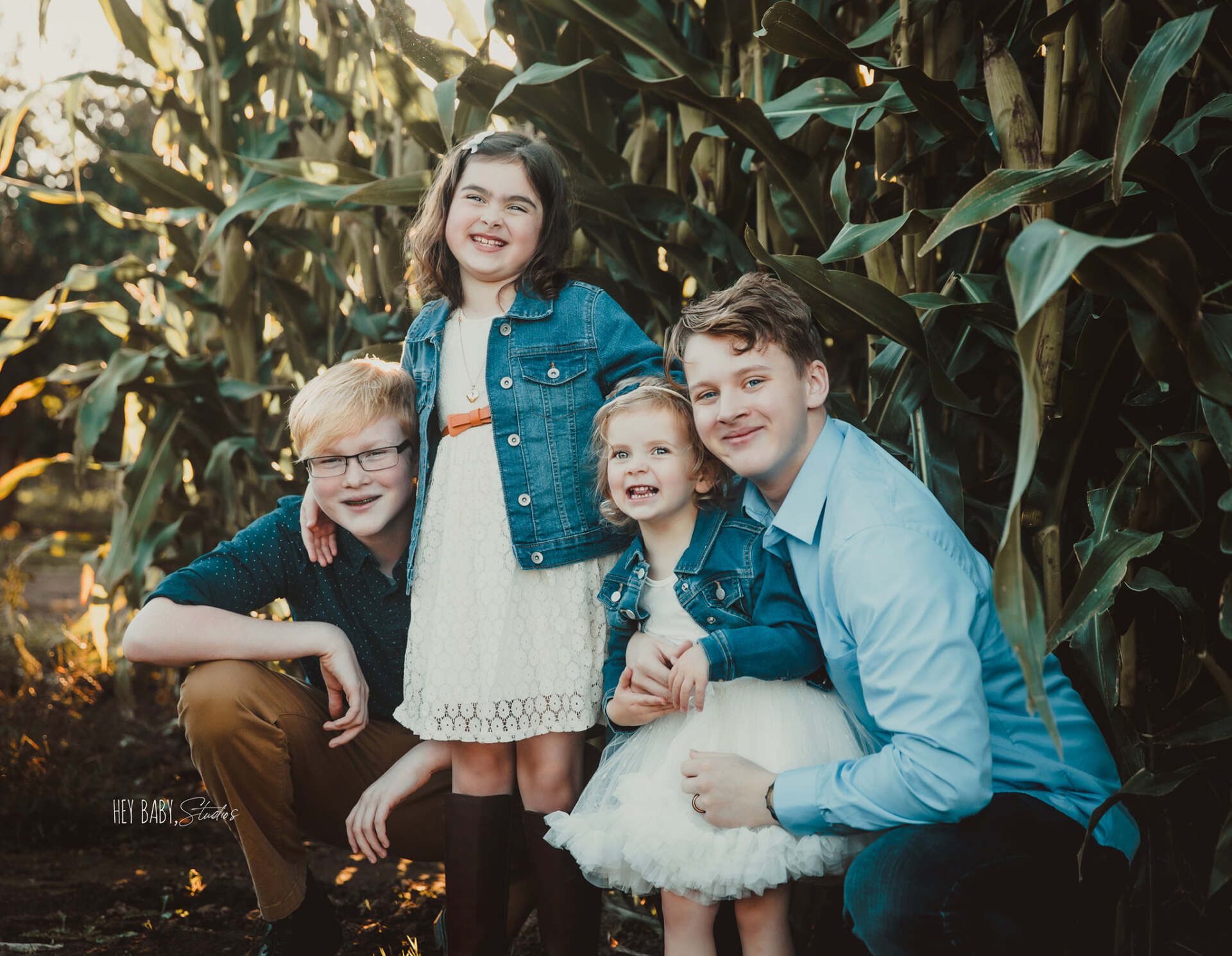 Family of four smiling, posing in front of corn stalks; denim jackets, light-colored dresses, autumn colors.