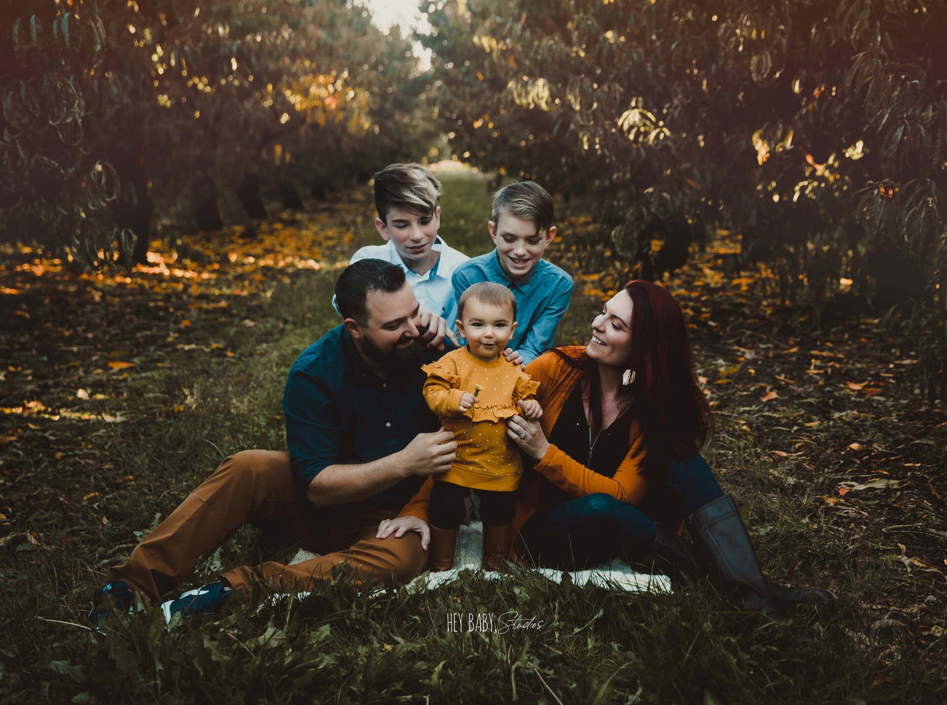 Family of five sitting on a blanket outdoors; two boys behind, parents flank toddler, all smiling.