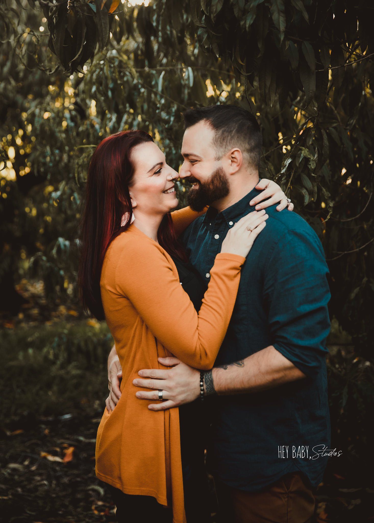 Couple embracing outdoors, smiling, woman in orange top, man in blue shirt, trees in background.