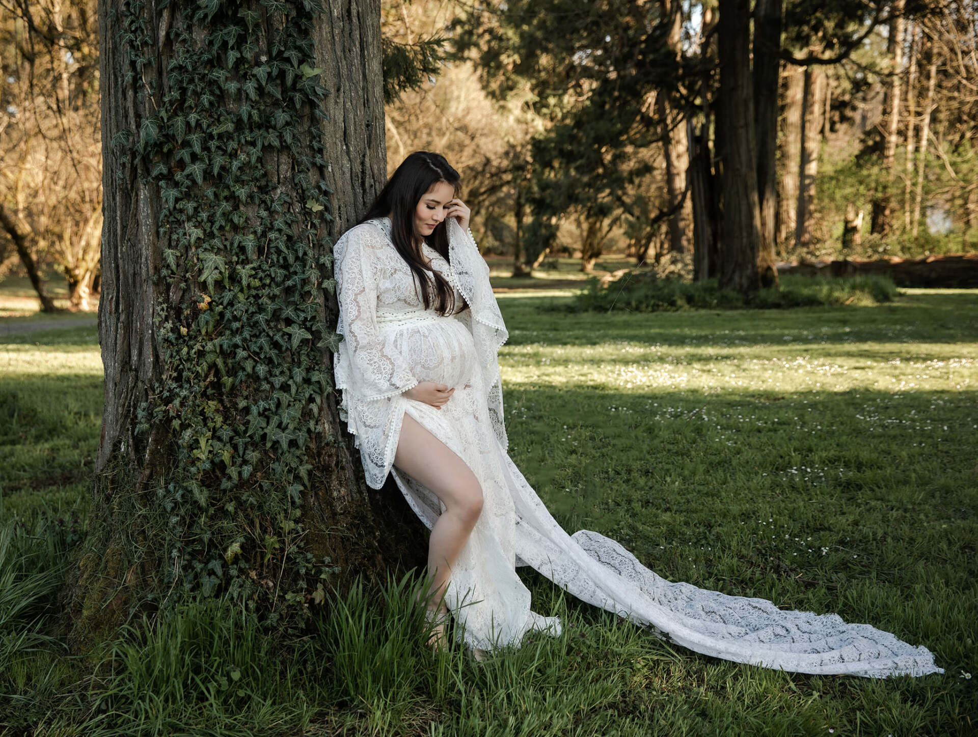 Pregnant person in white gown leans against a tree in a park, touching stomach.