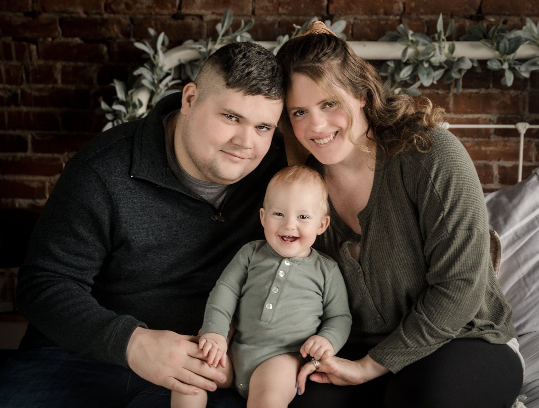 Family of three smiling, posing on a bed; brick wall background, decorated with greenery.