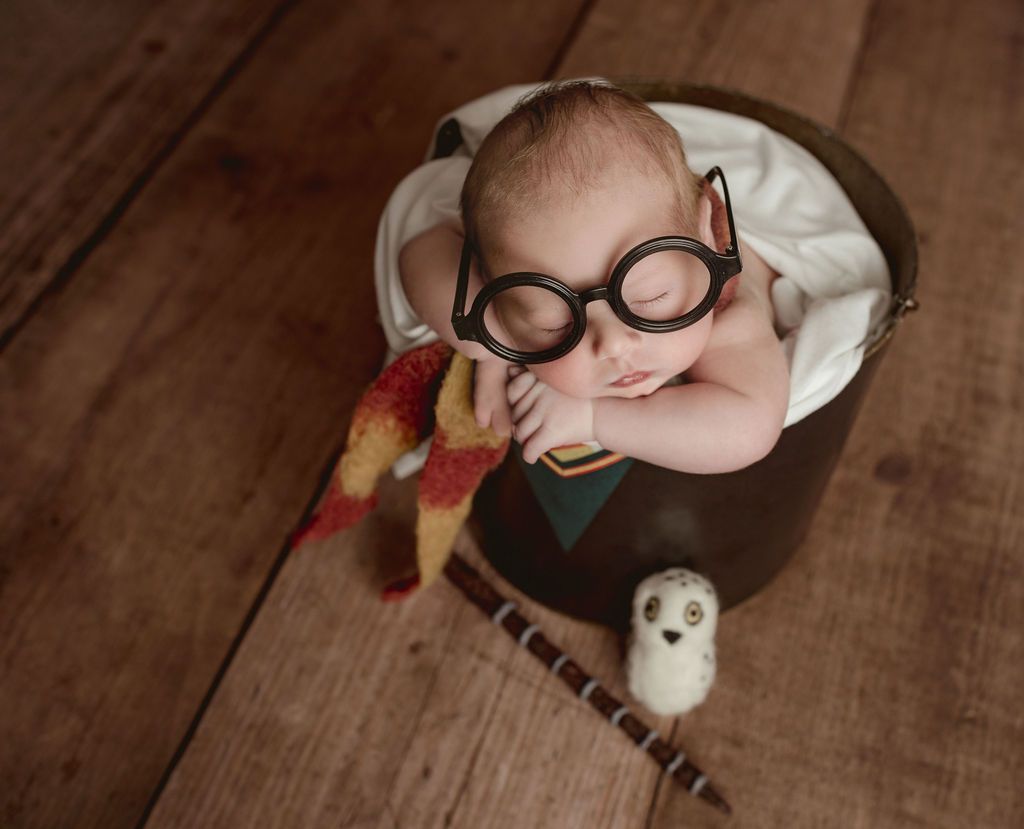 Newborn baby dressed as Harry Potter, wearing glasses and holding a wand, in a cauldron.