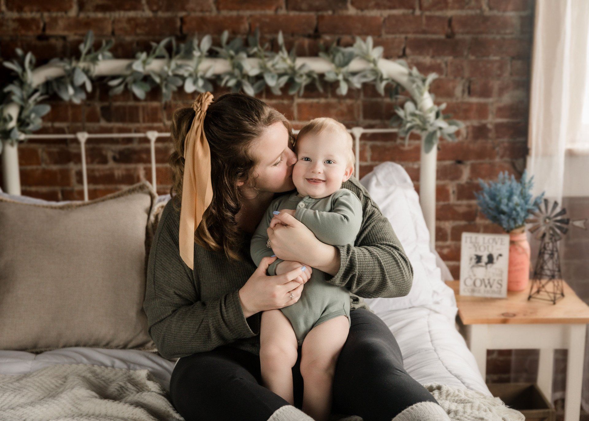 Woman kissing a baby on a bed with a brick wall backdrop.