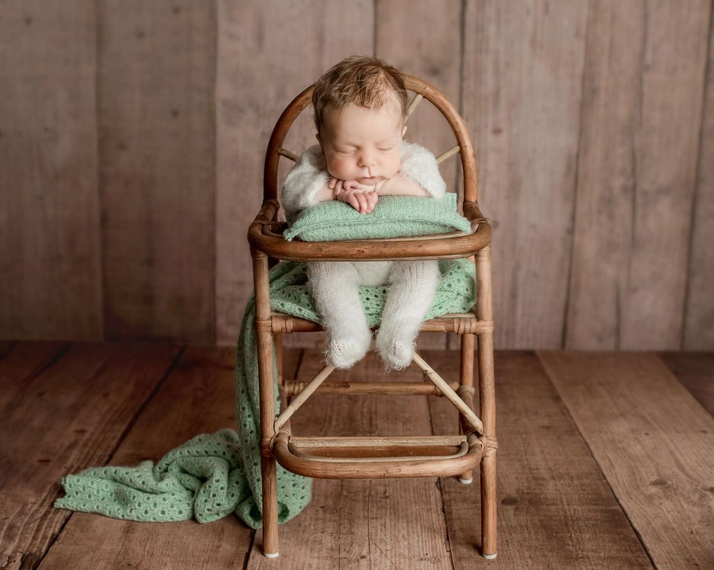 Baby asleep in a vintage wooden high chair, draped with a green blanket against a wood backdrop.