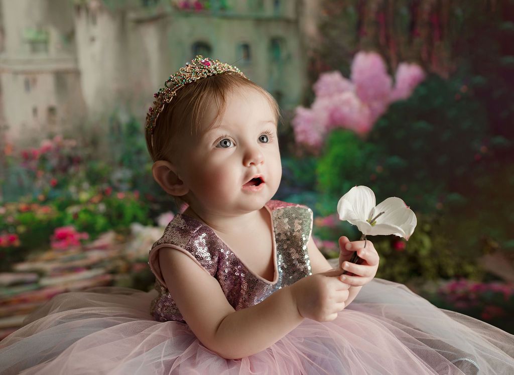 Baby in pink dress and crown, holding a white flower, against a fairytale backdrop.