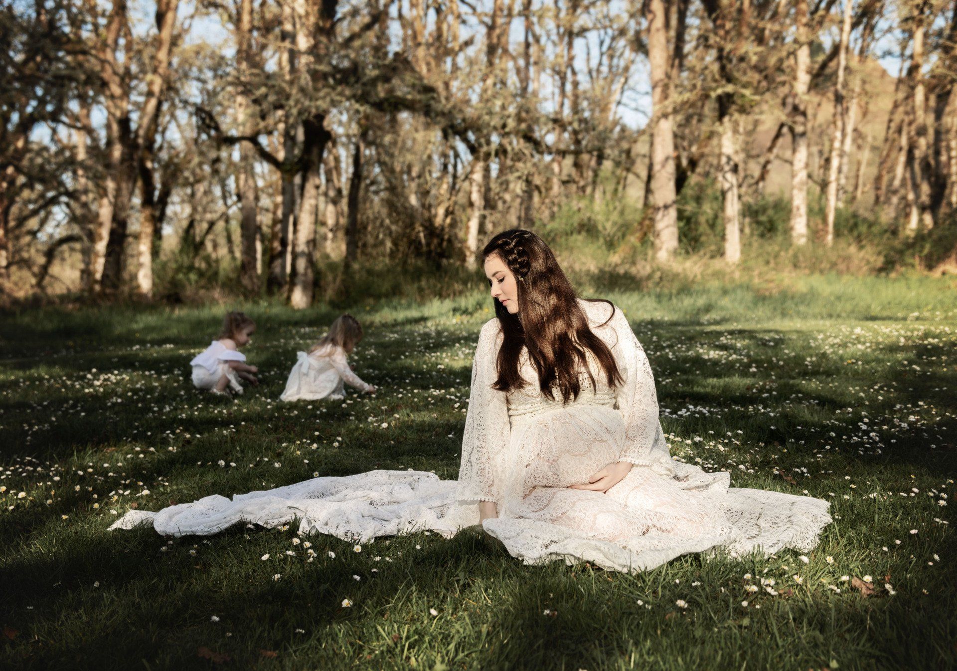 Woman in white dress sits in a field, two children play in the background, trees surround.