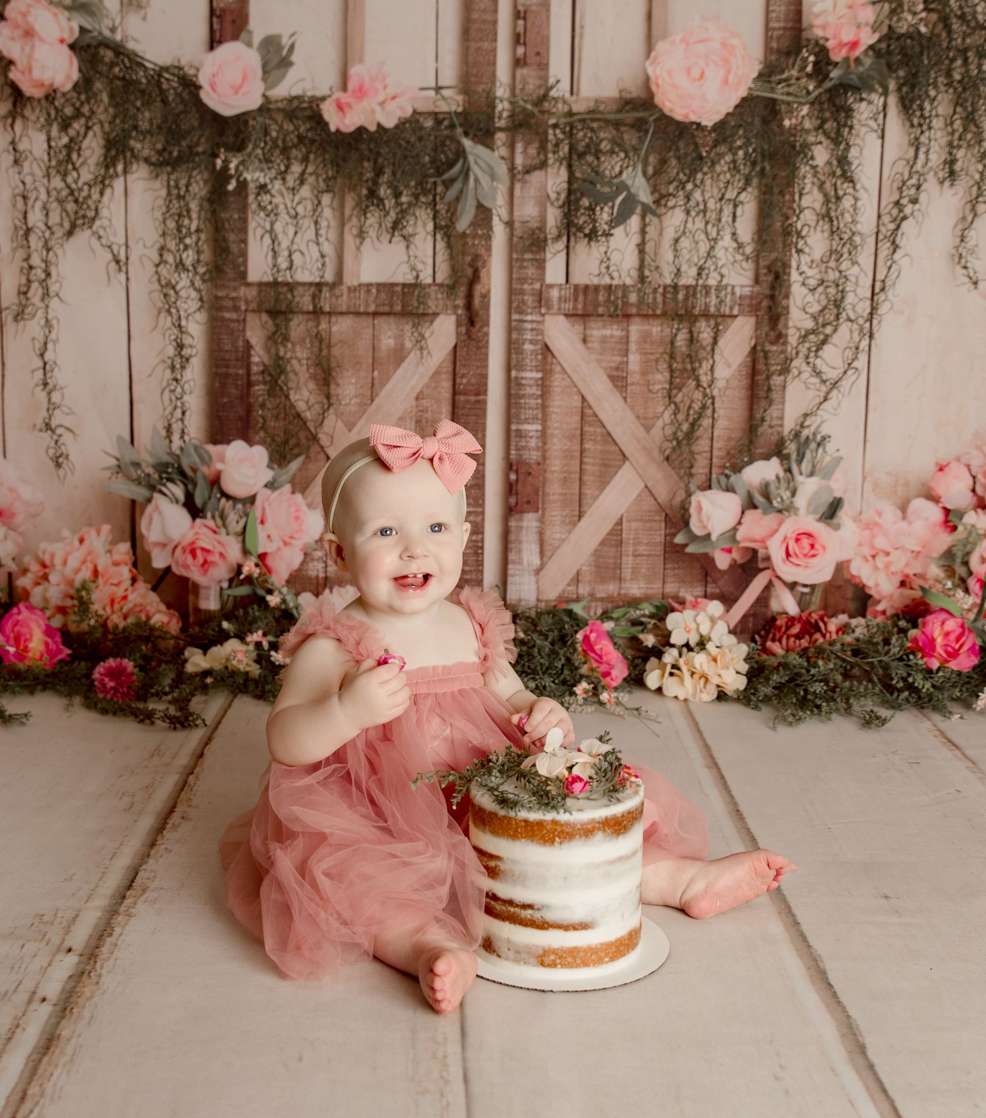 Smiling baby in pink dress and headband with a cake, sitting in front of a floral backdrop.