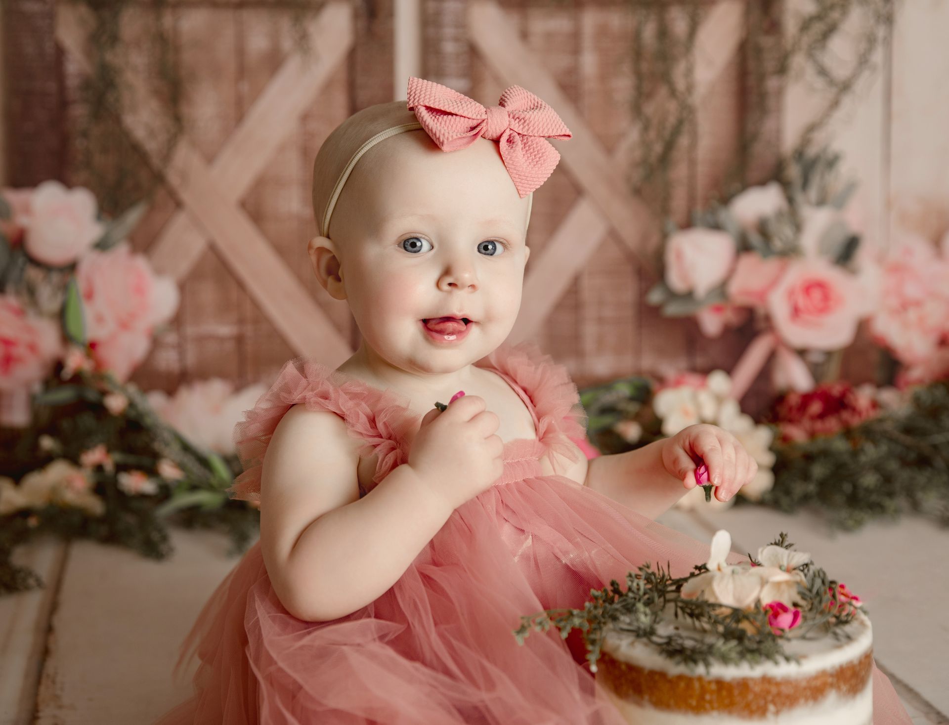 Baby in pink dress with a bow on head, smiling at cake with a candle in a floral setting.