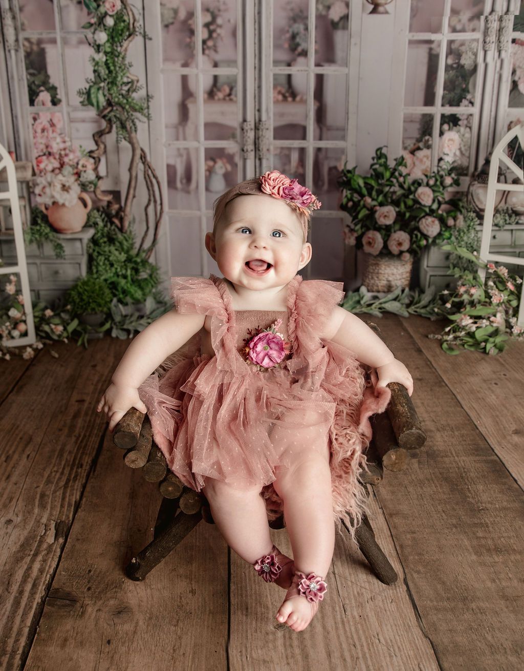 Baby in pink lace outfit smiles, sitting in a wooden chair, floral backdrop.