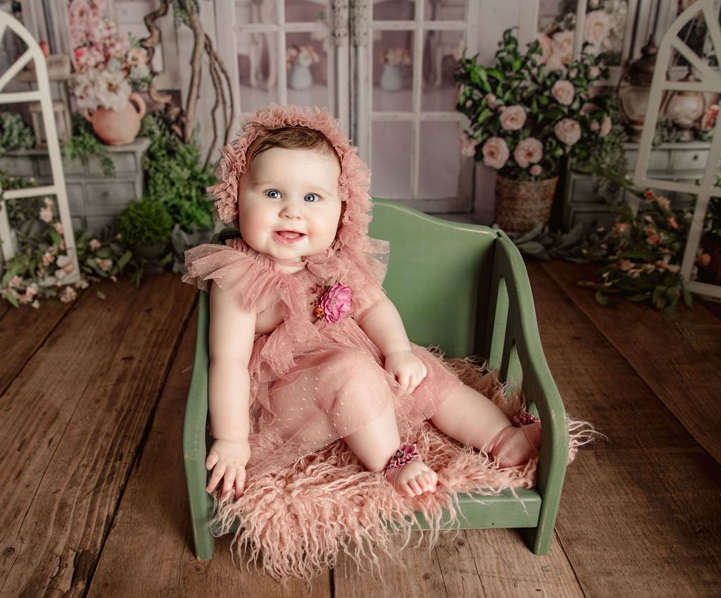 Smiling baby in a pink dress and bonnet sits in a small green bench, surrounded by flowers.